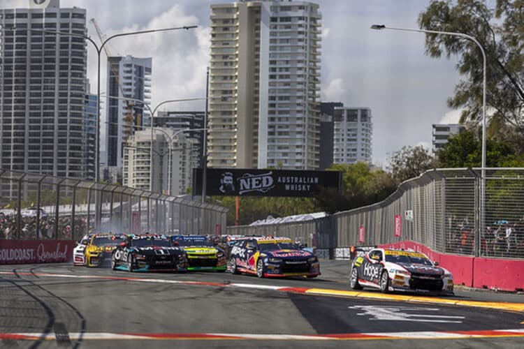 Race cars speed through a corner at the Boost Mobile Gold Coast 500 street circuit, with high-rise buildings and cheering spectators in the background.